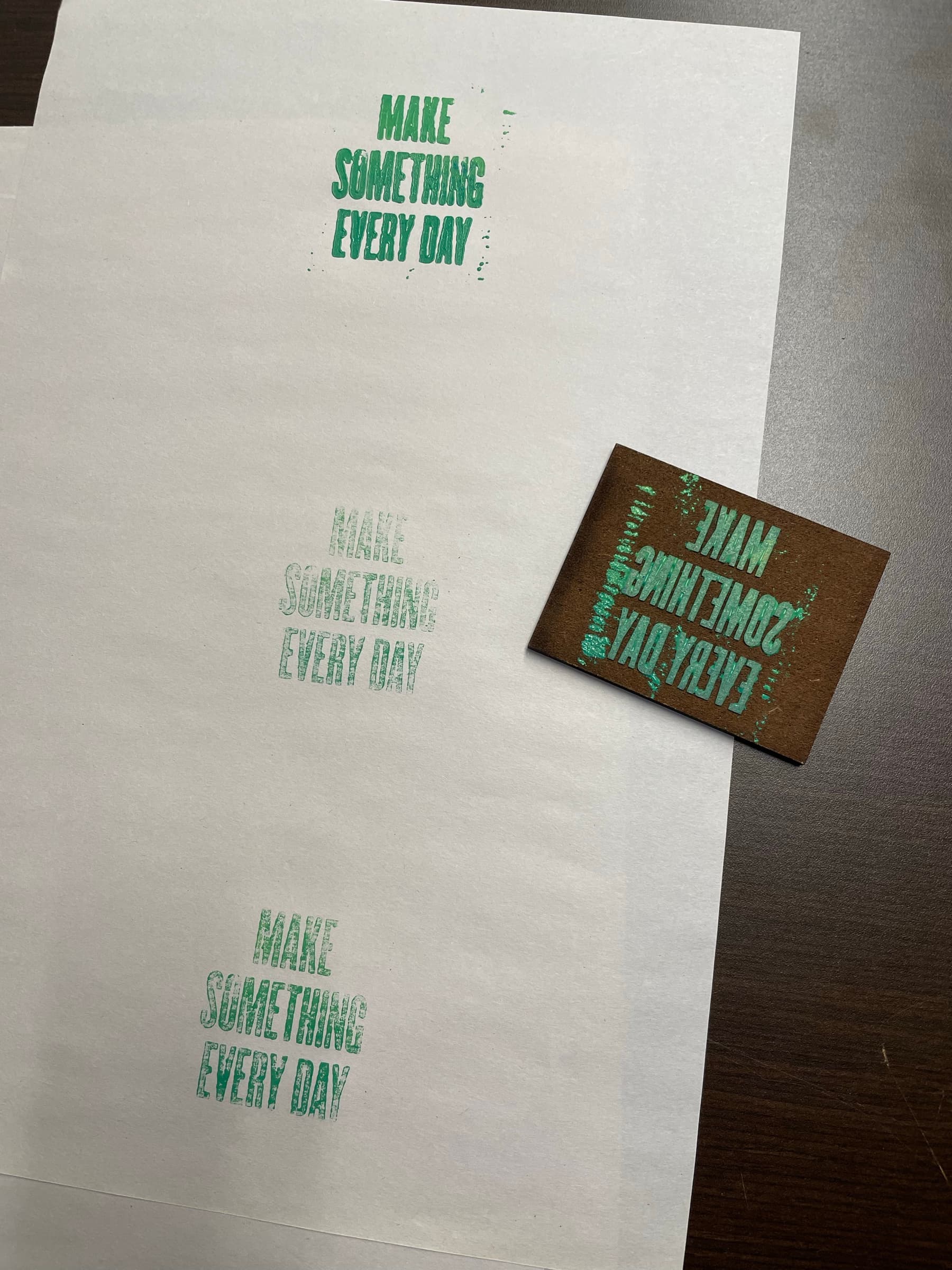A workshop flat-lay: the carved block reading MAKE SOMETHING EVERY DAY (mirrored) sits in the upper-third center on a brown background, with three pulls of the same phrase printed in green across a long sheet of cream paper below it, each pull progressively lighter as the ink loads off the block.
