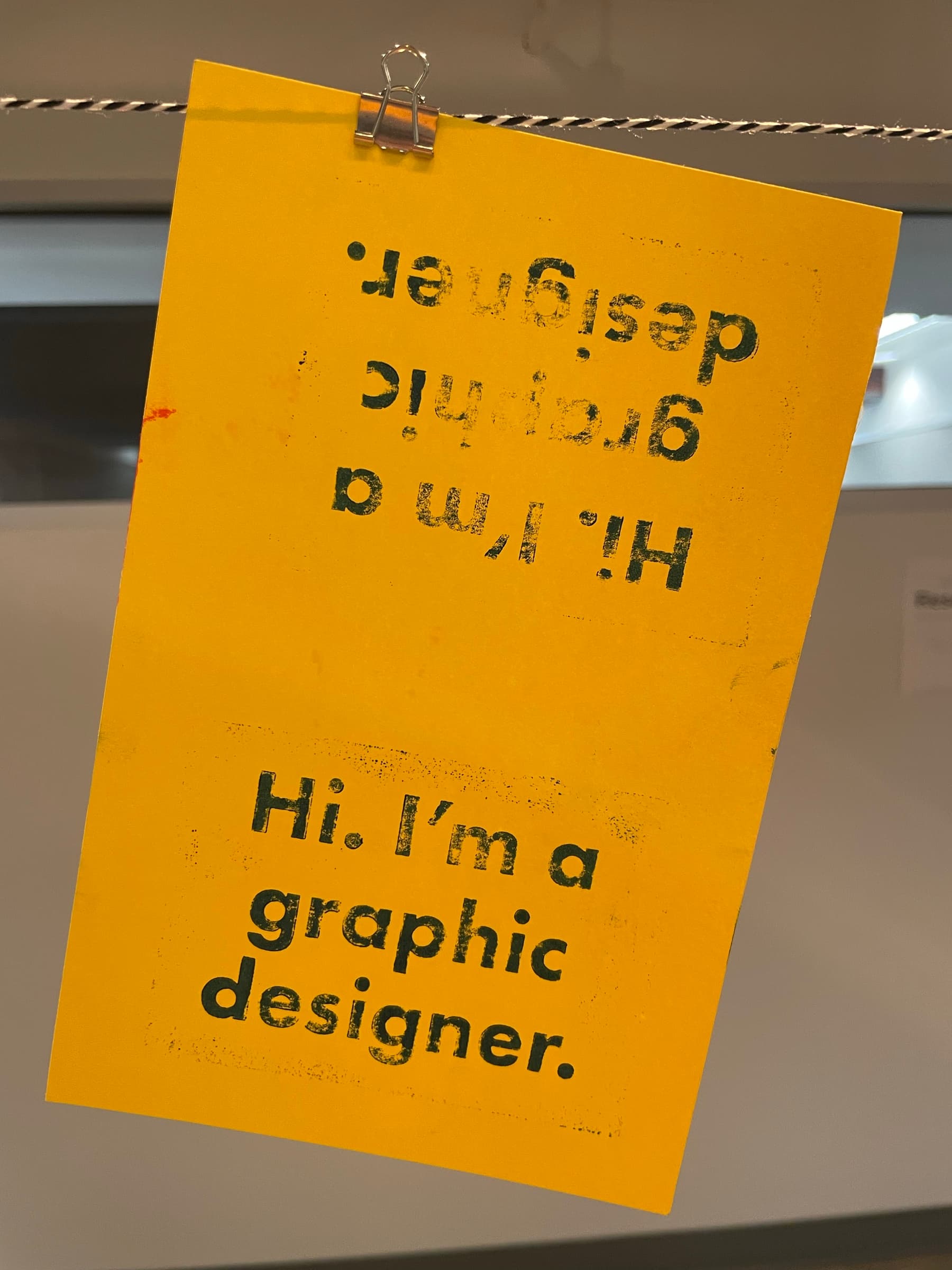 A hot-magenta-and-black hatched checkerboard print clipped to a clothesline by a binder clip in a studio space: a single small carved block stamped twenty-some times in tight registration to fill the sheet, alternating crosshatch directions to read as woven texture.