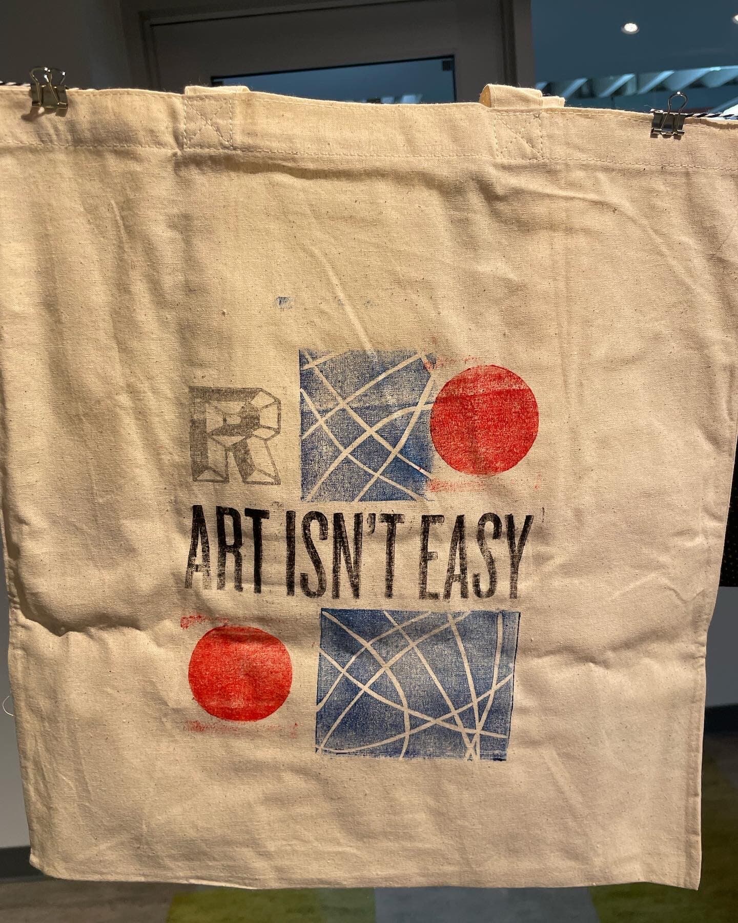 A natural canvas tote bag hung from clips against a white wall: ART ISN'T EASY set in chunky distressed black display caps in the lower-third center, with two indigo carved-line bramble blocks framing the type on either side and two scarlet ink discs anchoring the upper-left and lower-right corners as accent marks.