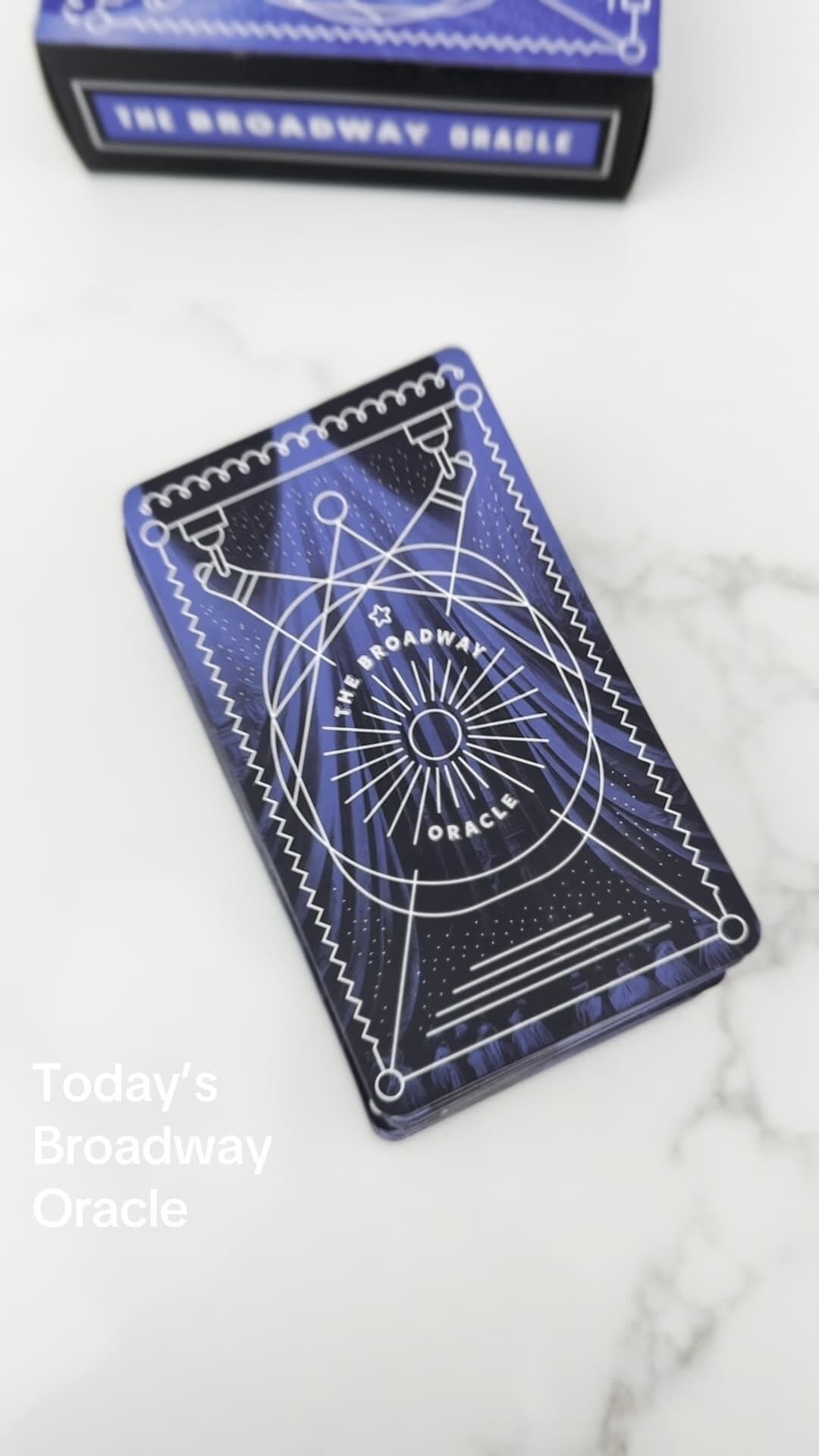 The Broadway Oracle deck on white marble: a black-and-royal-blue tuck box at the top of the frame with the THE BROADWAY ORACLE wordmark on the side, and a deck of cards splayed on the marble below. Each card shows a royal-blue back with white linework: twin theatre spotlights, a stage curtain, a sun-ray oracle emblem reading THE BROADWAY ORACLE, and zigzag borders that read as marquee bunting.