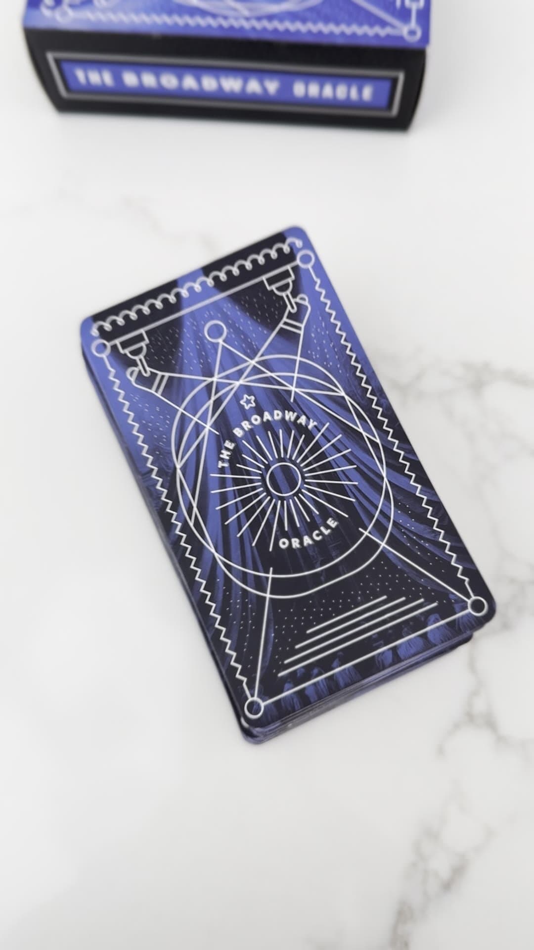 An overhead frame from a daily reel: a hand fanning the deck on white marble, the box of the deck visible in the lower-left corner, the caption 'Today's Broadway Oracle' set in white sans on the lower edge of the frame.