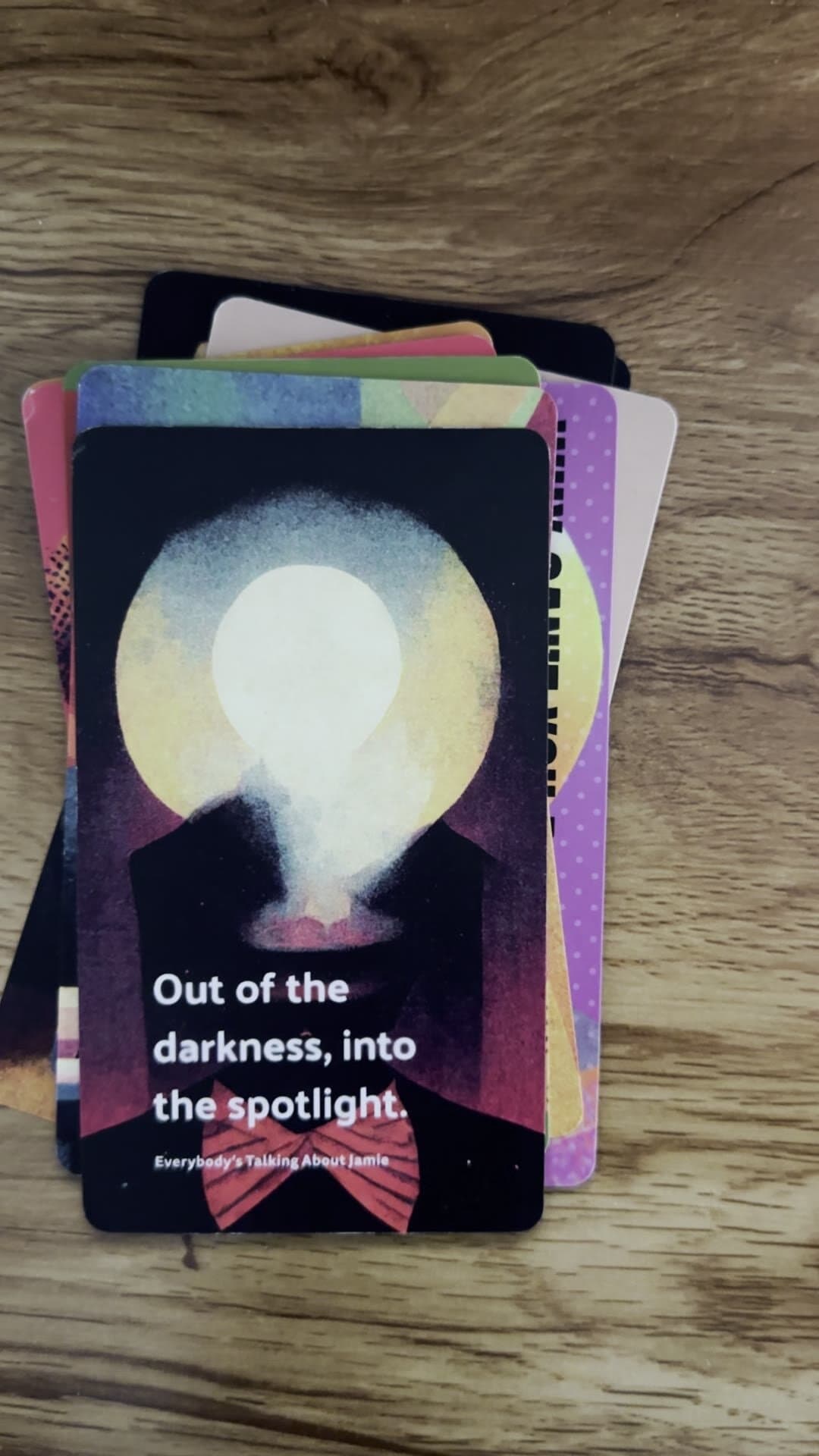A printed card face: an illustrated figure in a tuxedo with a glowing yellow-and-pink lightbulb where its head should be, against a black ground. 'Out of the darkness, into the spotlight.' set in white sans body copy across the lower half. 'Everybody's Talking About Jamie' as the small white footer citation.