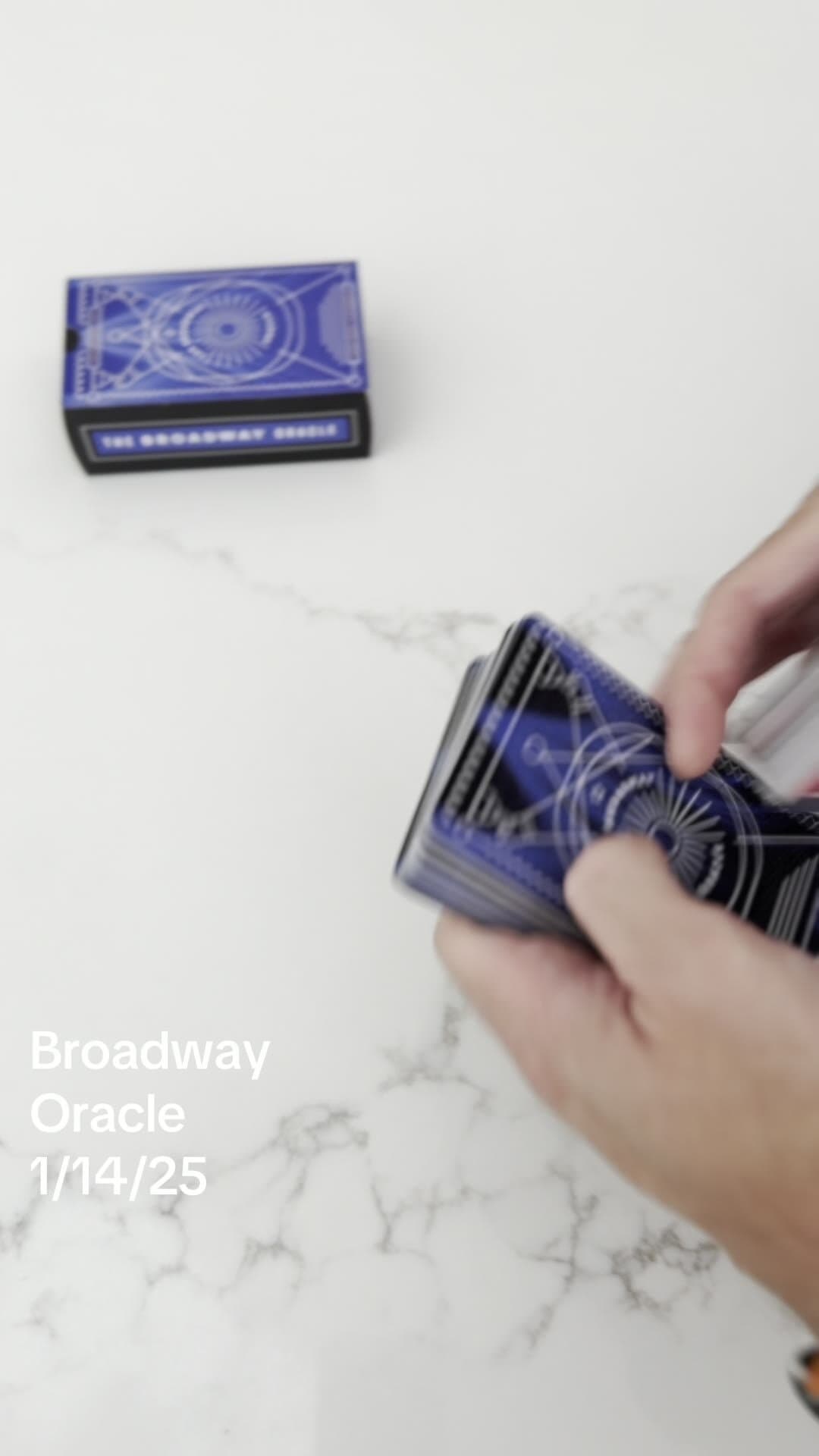 Another reel frame: the closed Broadway Oracle box sitting on the marble in the upper-left, hands shuffling cards in the lower-right, with 'Broadway Oracle 1/14/25' set in white sans across the lower-left of the frame as a date stamp for the daily pull.