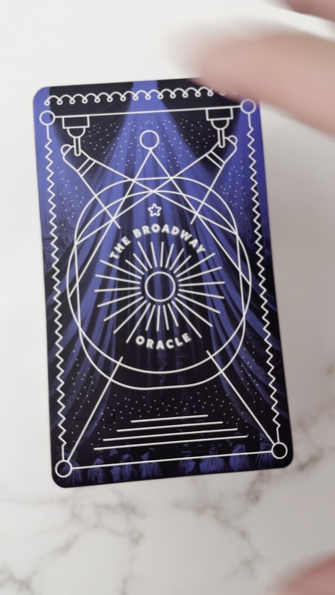 A single printed card held to the camera: same composition rendered in white-on-royal-blue, the curtain folds painted into the negative space behind the spotlights, the sun-ray oracle emblem centered, the printed dimensions and proportions of an actual playing card.