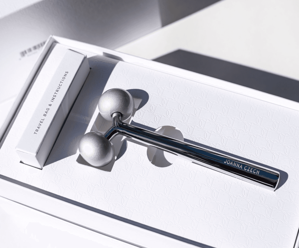 A close-up of the polished steel facial massager emerging from the white box, JOANNA CZECH engraved in small caps along the handle, the metal catching the light against the white field