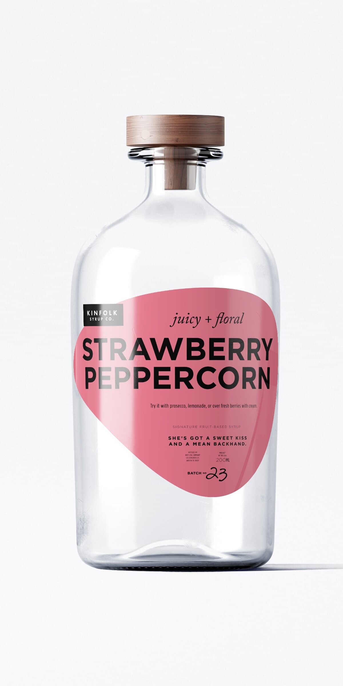 The Strawberry Peppercorn bottle: same apothecary glass and wooden cap, hot dusty-pink teardrop label with KINFOLK SYRUP CO. tag in the upper-left.