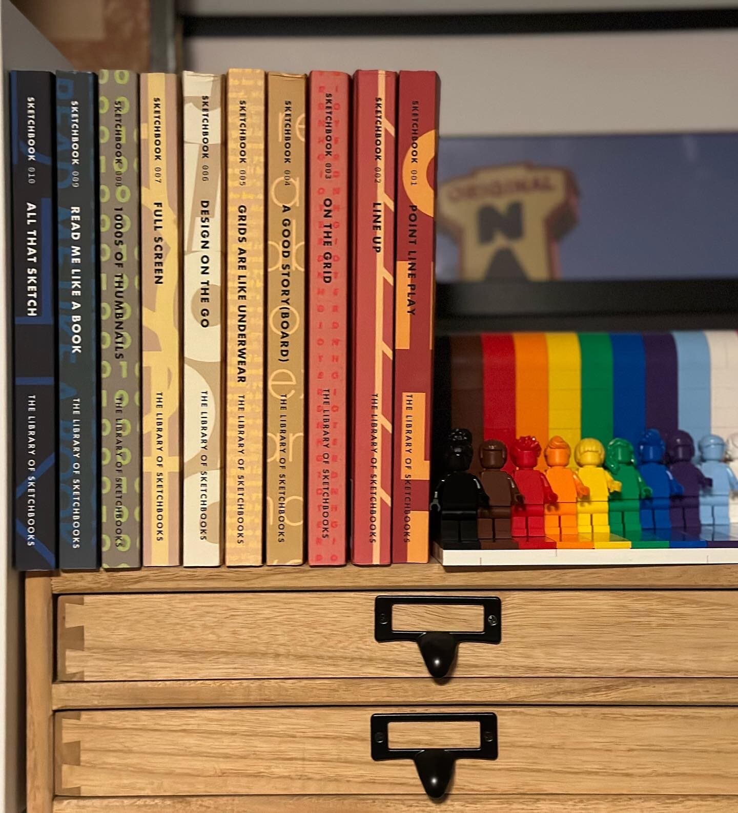 A row of nine sketchbooks standing upright on a wooden cabinet, each one a different cover color (deep navy, teal, gold, cream, peach, scarlet) with the volume number and title printed on the spine. THE LIBRARY OF SKETCHBOOKS runs across all the spines in a uniform display caps. A row of rainbow Lego figures stands beside them as bookend.