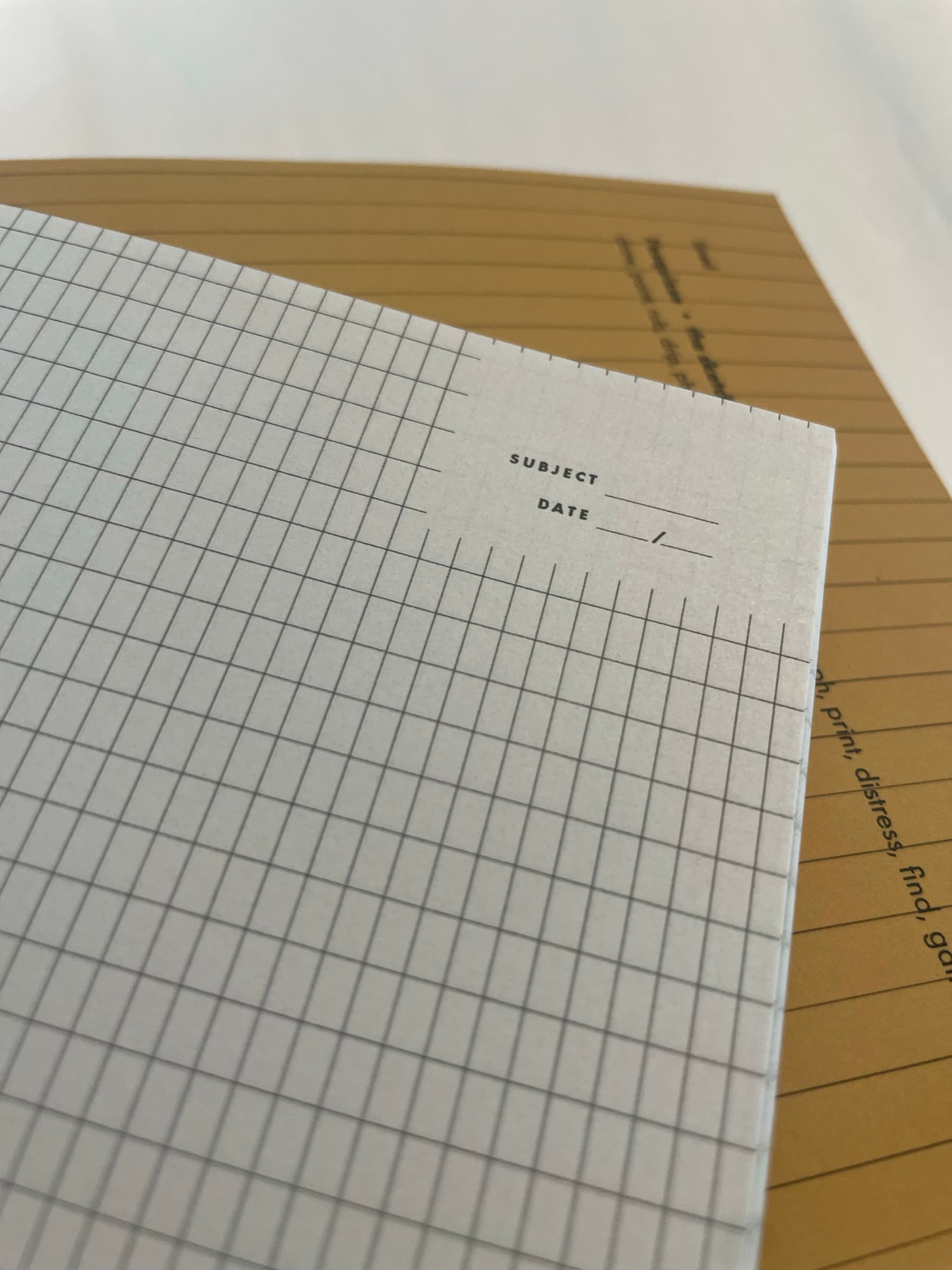 A close-up detail of one of the page templates: a SUBJECT and DATE header in tiny tracked caps in the upper-left of a fine square grid. The kraft endpaper of the book itself shows underneath, with 'rub, drip, photograph, print, distress, find, gather' partially visible in the gutter type.