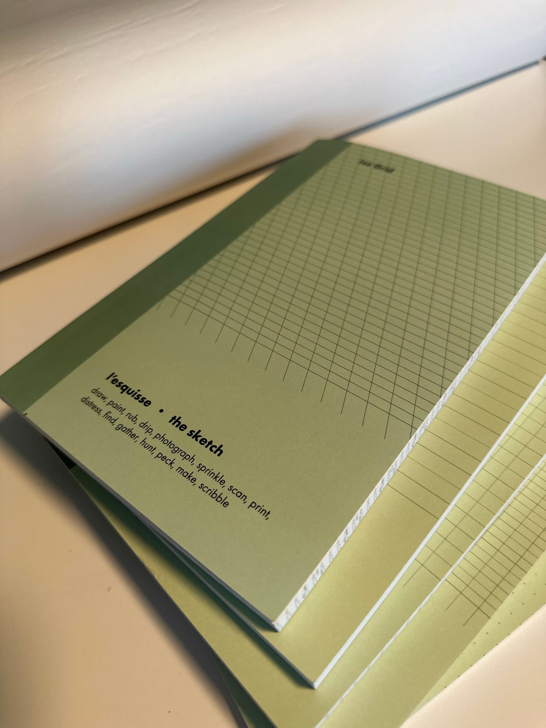 An interior page of another volume: a soft-green ground with a fine grid, the page-header reading 'iso grid / l'esquisse · the sketch / draw, paint, rub, drip, photograph, sprinkle, scan, print, distress, find, gather, hunt, peck, make, scribble' set in tracked sans body copy along the upper edge of the spread.
