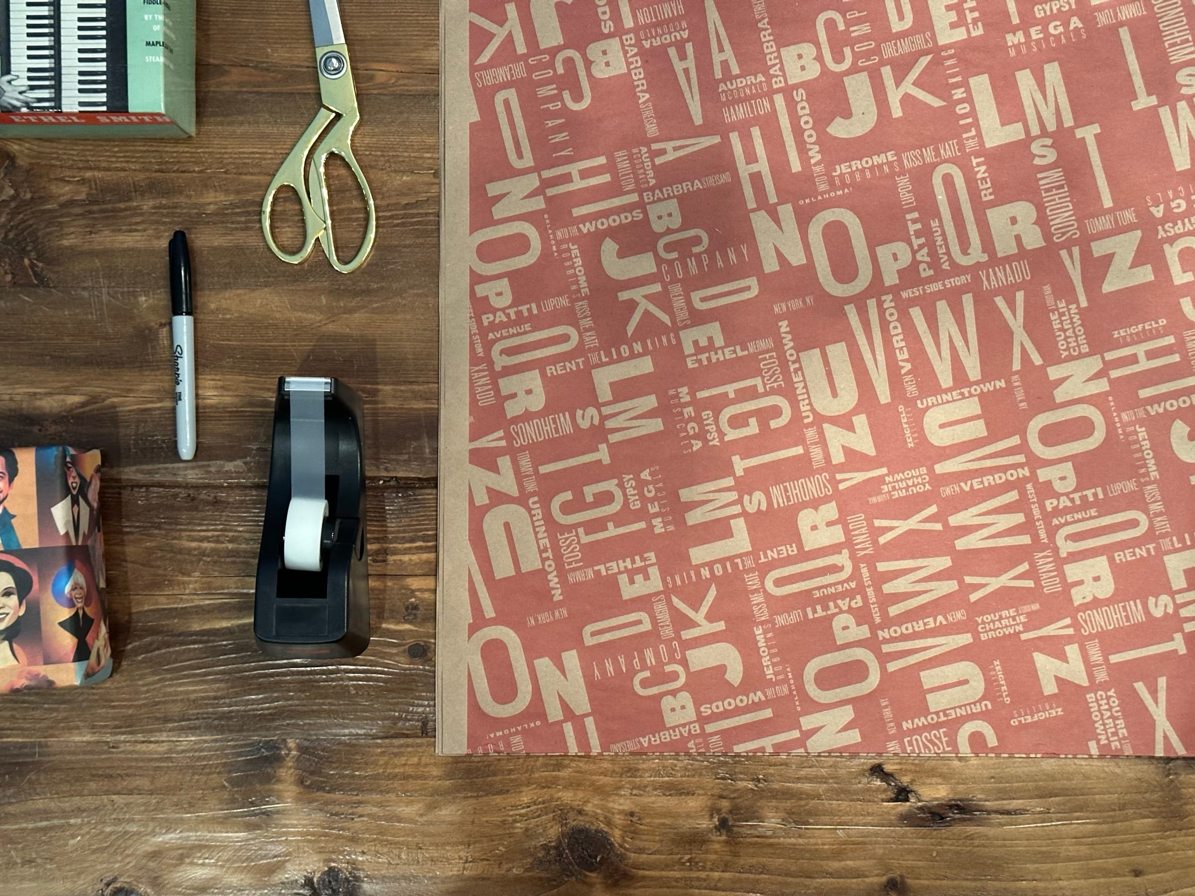 An overhead view on a dark wood table: a sheet of coral wrapping paper rolled out across the right two-thirds of the frame, printed in cream with a dense typographic pattern of letters and Broadway show titles. A black tape dispenser, gold-handled fabric scissors, and a Sharpie sit at the edge of the sheet, ready to wrap something.