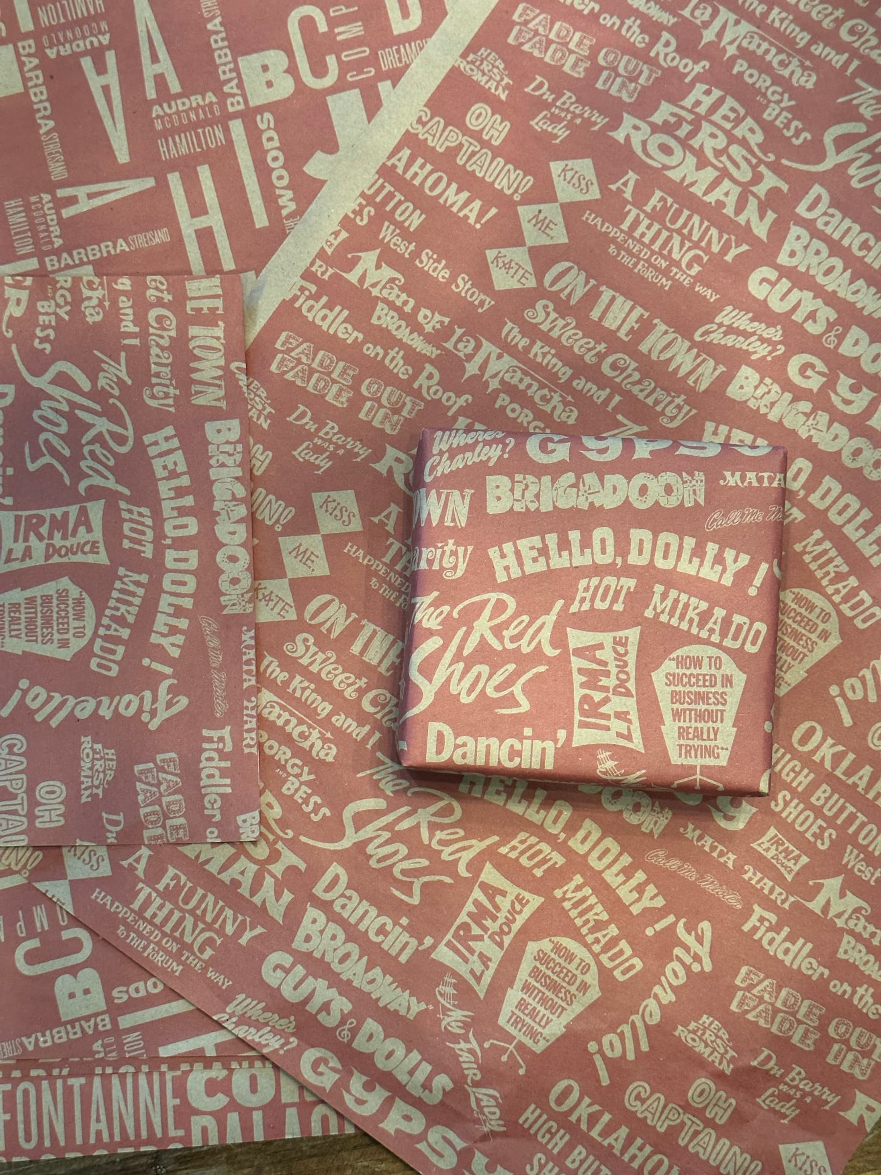 An overhead detail of the alphabet paper with a small wrapped gift box sitting on top of the sheet. The same Hello Dolly!, Brigadoon, Guys & Dolls, Oklahoma!, A Funny Thing, On the Town, Captains, Mata Hari pattern wrapping the box, and the rest of the sheet pattern visible as the ground around it.