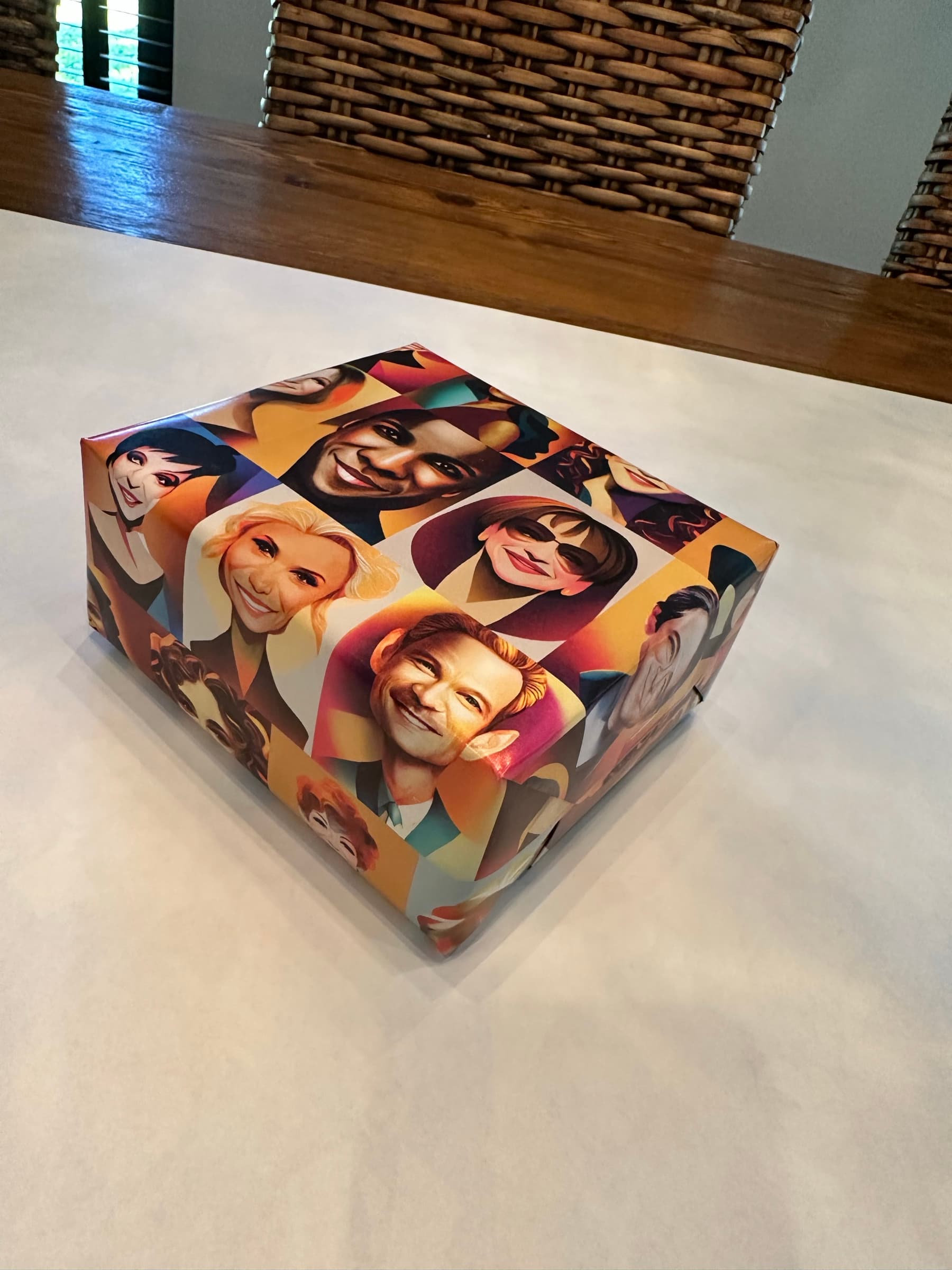 A small gift box wrapped in the portrait paper, photographed from above sitting on a tile floor: nine portraits visible across the front of the box, the wrapping crease catching directional sunlight.