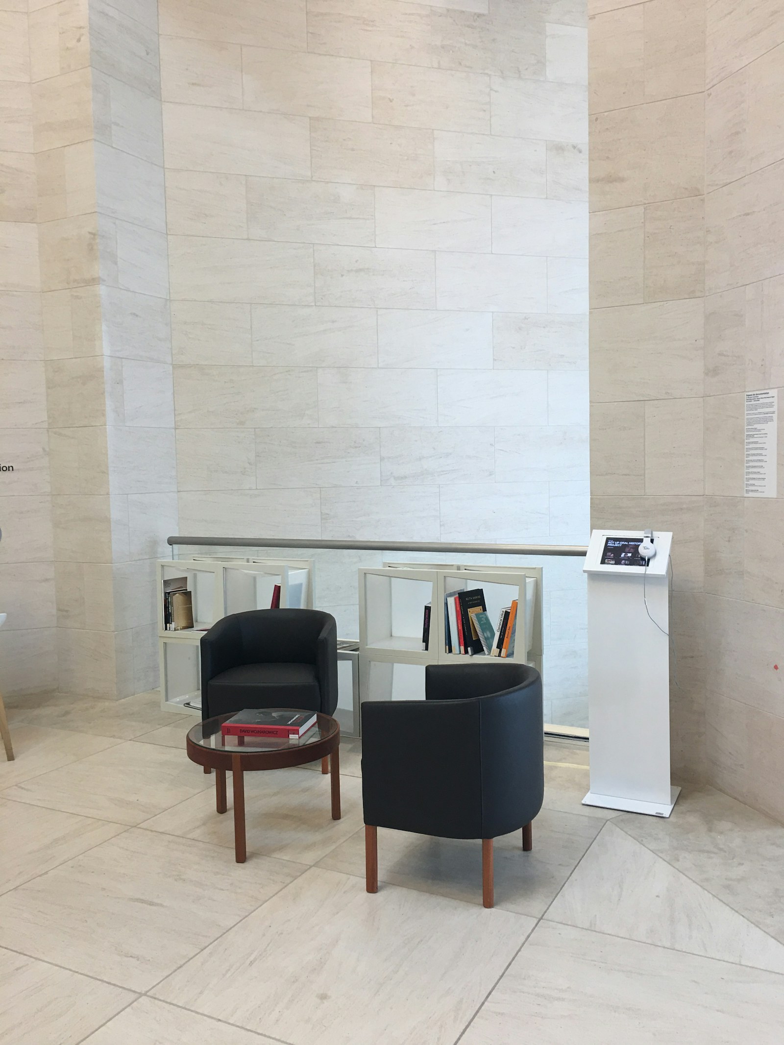 A minimal museum interior: pale gridded tile walls, two black tub chairs, a small podium